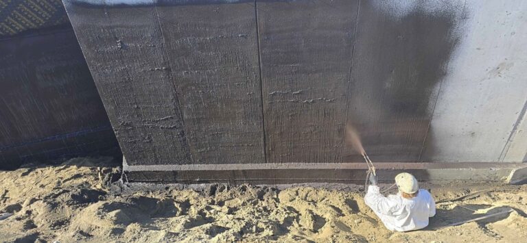 Crew member spraying waterproofing along the base of a long foundation wall