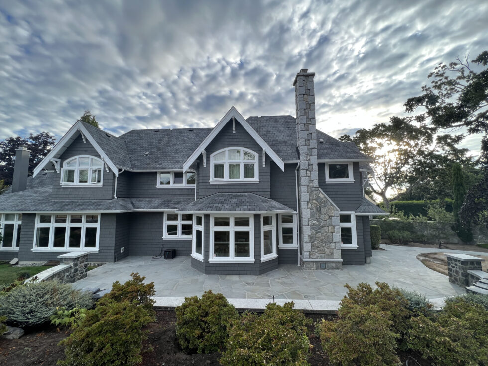 Wide flagstone patio behind a grey shingle-style home with a tall stone chimney