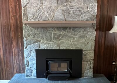 Large natural stone fireplace featuring a rustic wood mantel and a black wood-burning insert, installed in a residential home in Victoria, BC.