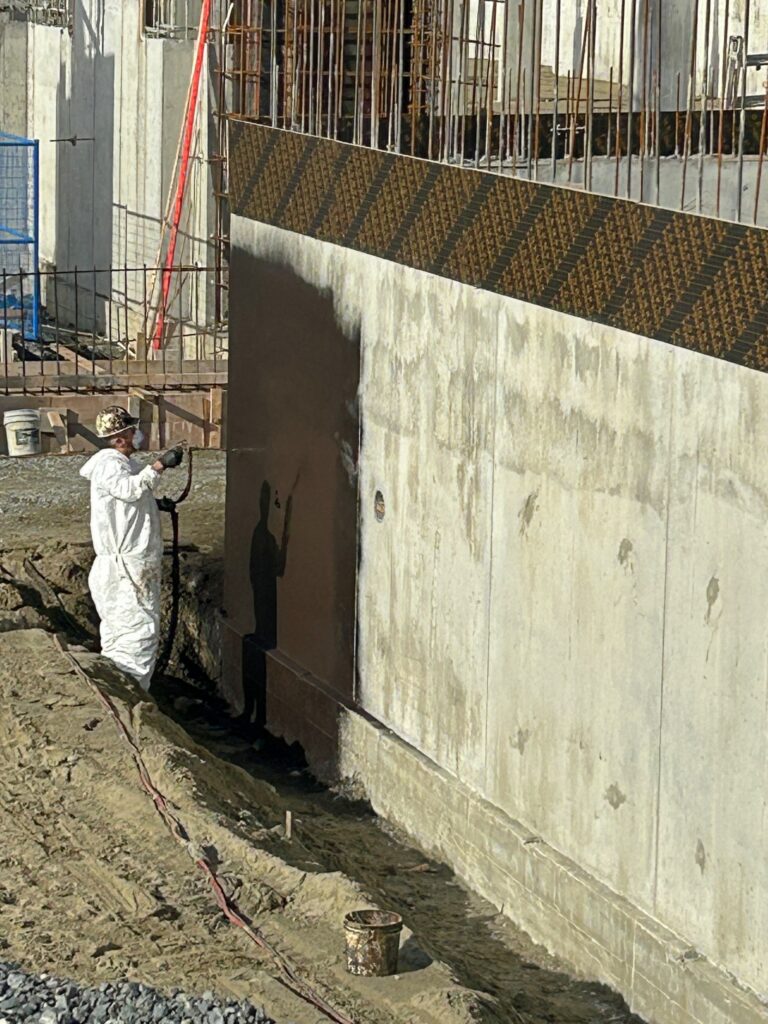Crew member in a protective suit beside a freshly waterproofed foundation wall