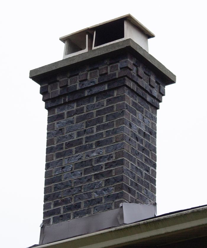 Dark blue-grey brick chimney with a corbelled top, concrete crown and cap