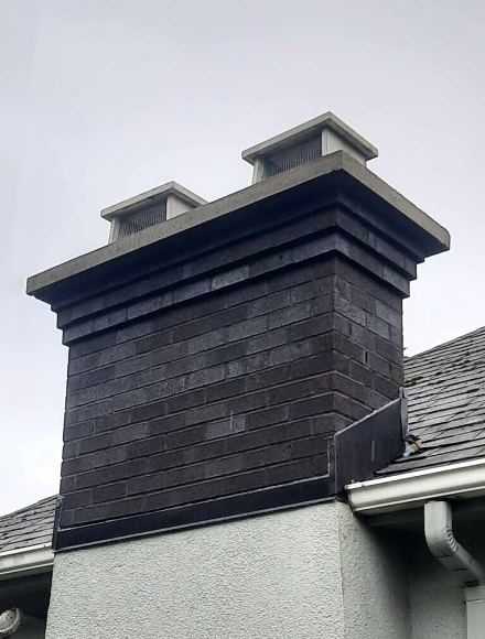 Dark charcoal brick chimney with a concrete crown and two caps above a grey stucco home