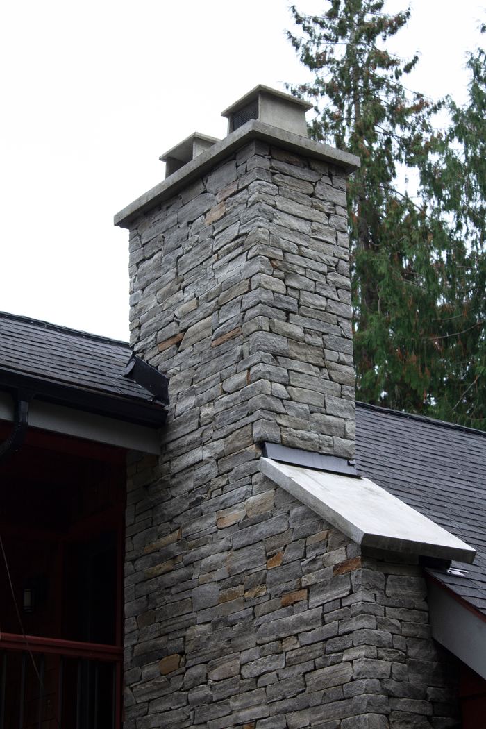 Grey ledgestone chimney with a metal cricket where it meets the roof and twin caps