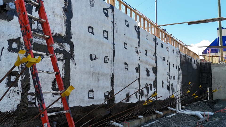 Foundation wall covered in fastened drain mat beside a ladder