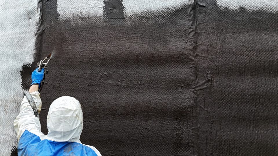 Close-up of a crew member in a protective suit spraying black waterproofing membrane